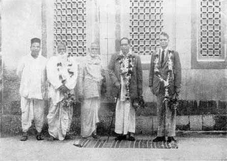 Five men standing outdoors for a formal photograph, two in suits with garlands and one in simple traditional cloth.
