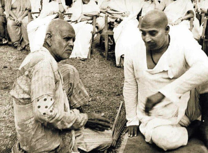 Sant Gadge Baba seated on the ground in patched clothes, facing Rashtrasant Tukdoji Maharaj, with a broom between them.
