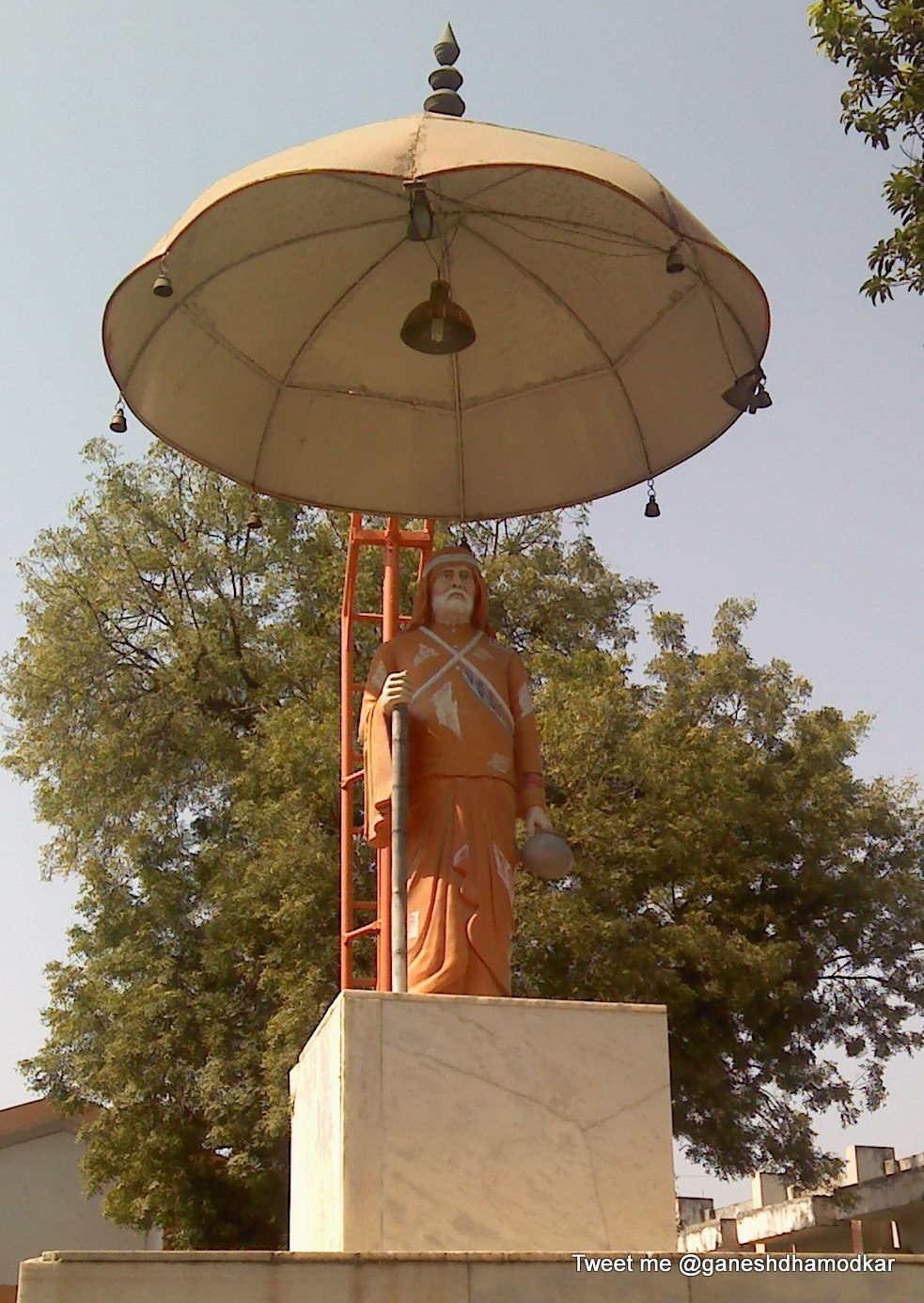 Painted standing statue of a saint under a ceremonial umbrella, holding a staff and a small pot, at Nagpur.